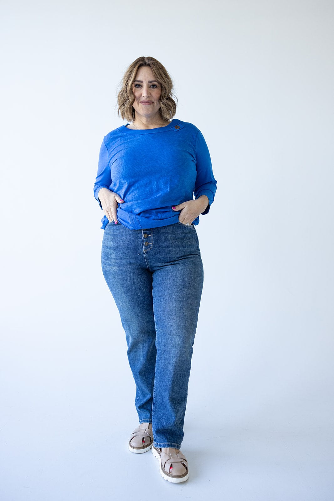A person wearing a blue long-sleeve shirt and high-rise jeans stands against a plain white background.