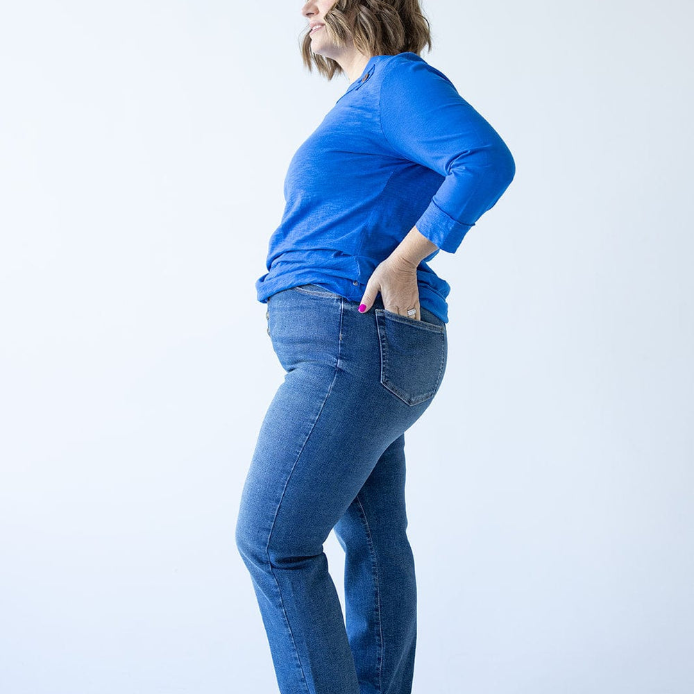 Woman in a blue sweater and high rise jeans standing sideways with hands in back pockets against a light background.