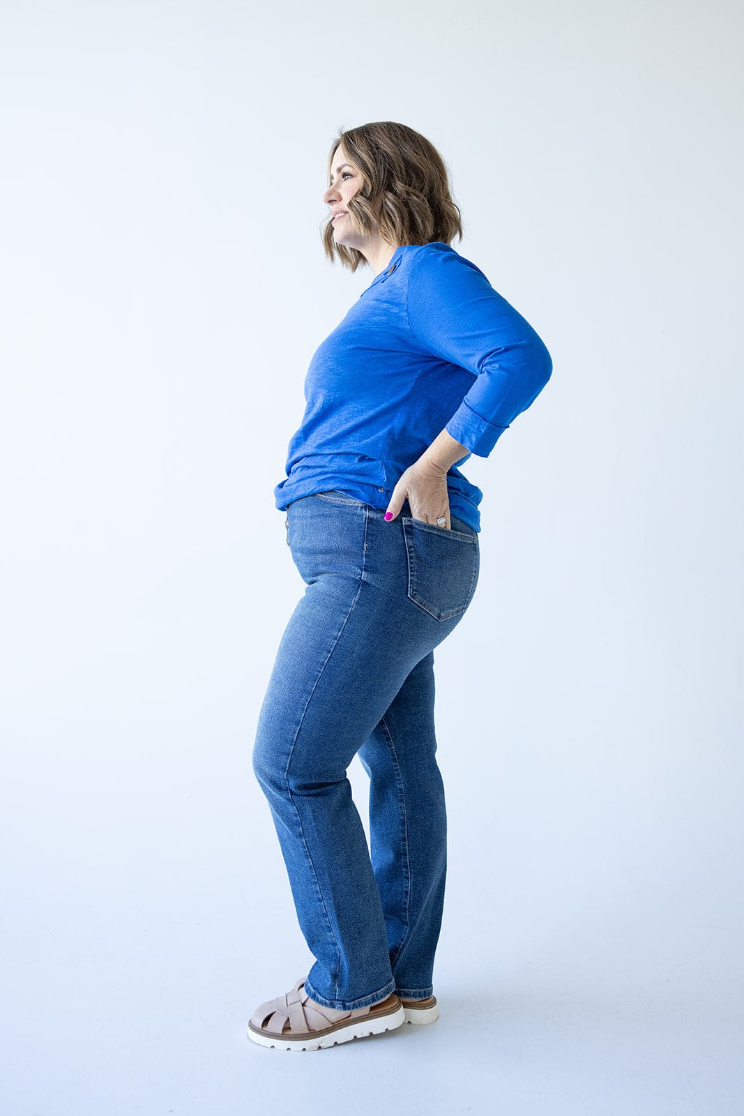 Woman in a blue sweater and high rise jeans standing sideways with hands in back pockets against a light background.