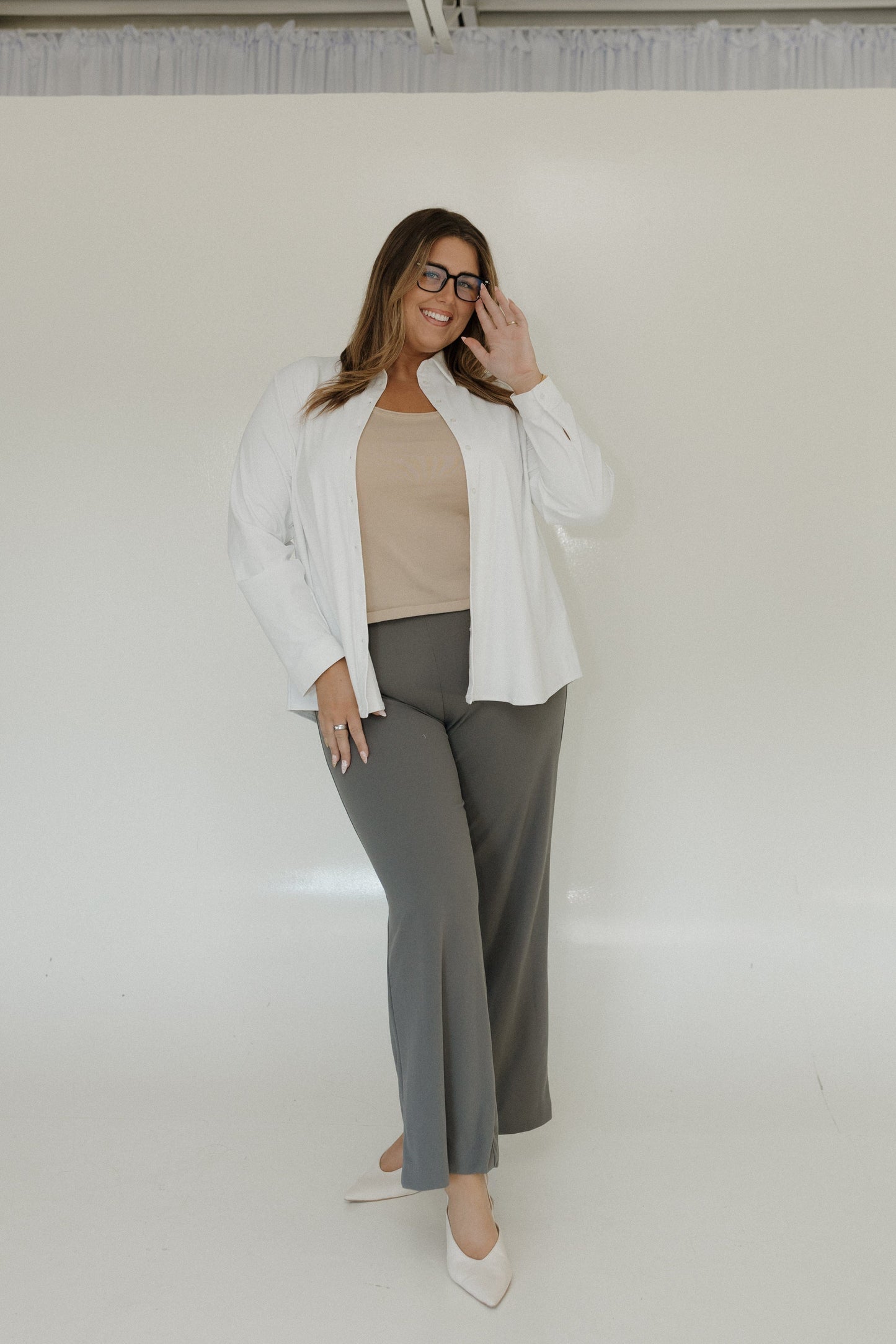 Woman wearing a white shirt, beige top, gray pants, and white shoes against a plain background