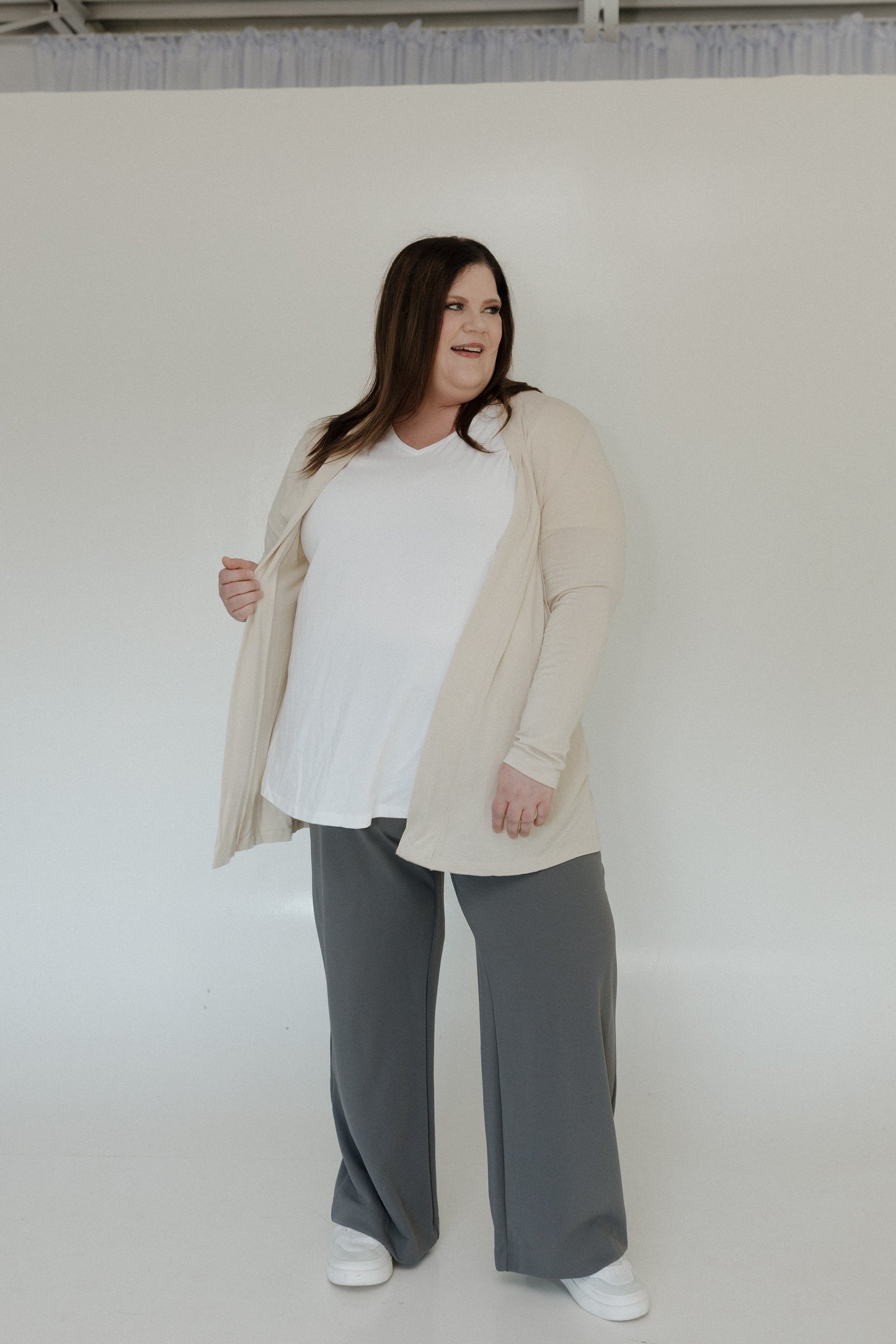 Woman wearing a beige cardigan, white shirt, and gray pants against a plain background