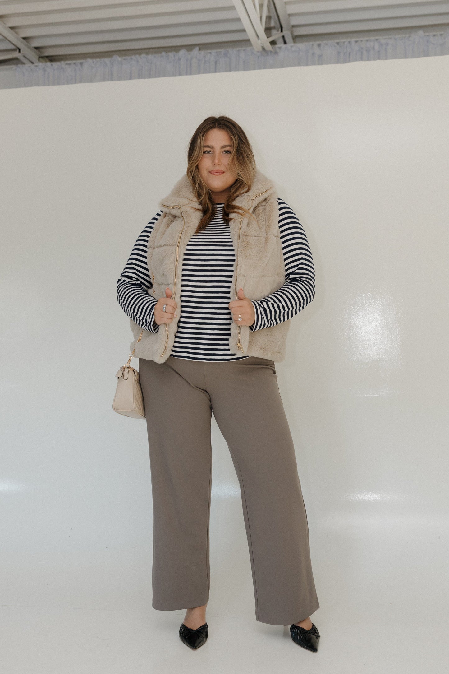 Woman wearing a striped sweater, beige vest, and beige pants against a white background