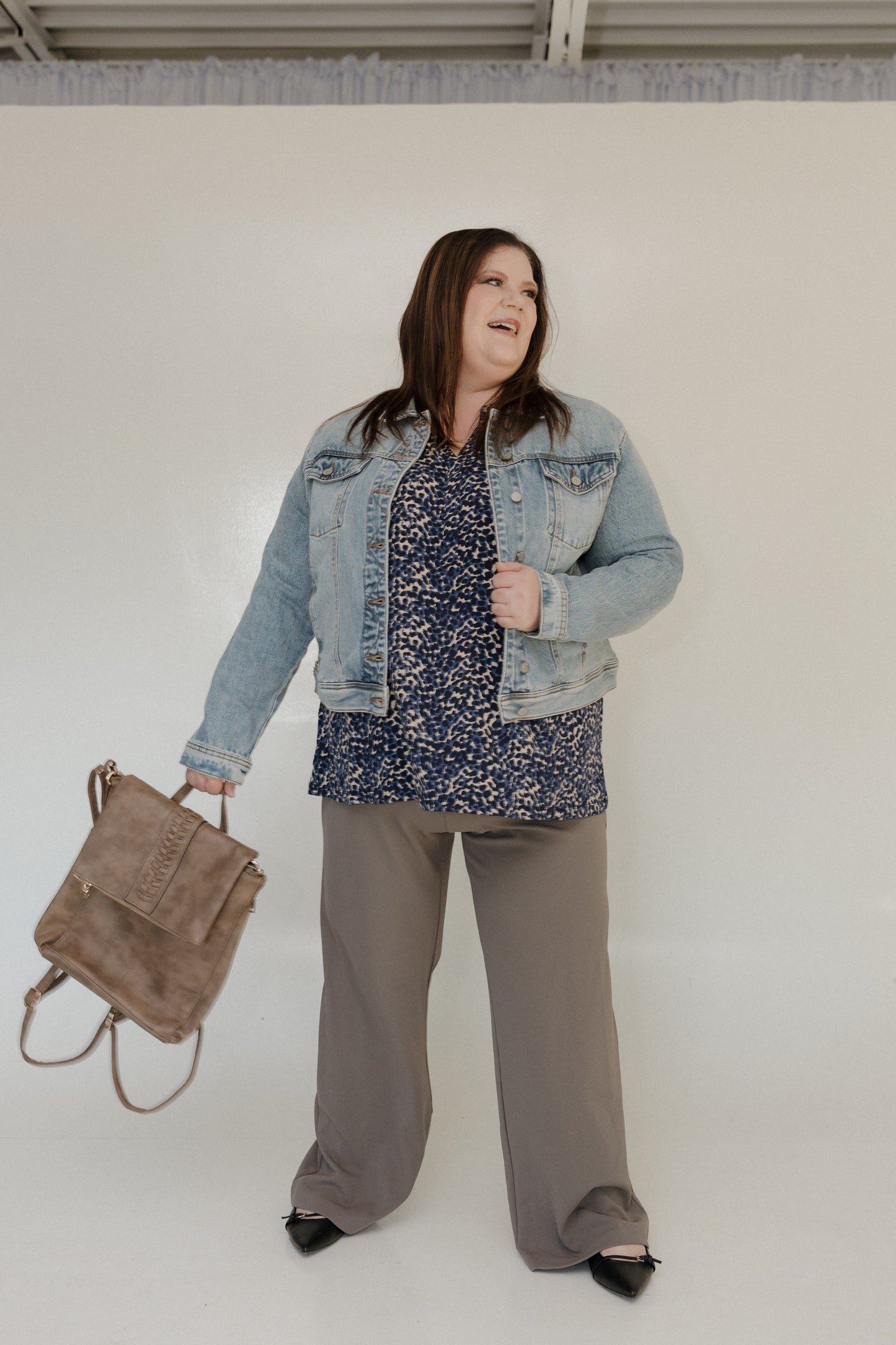 Woman wearing a denim jacket, floral blouse, and gray pants holding a brown bag against a white background