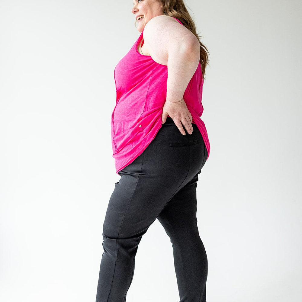 A woman in a pink top and black pants, with a slim straight leg design, stands sideways, smiling confidently with one hand on her hip in a bright room.