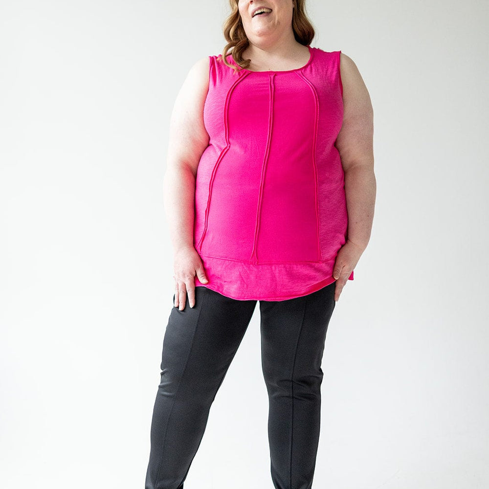 A woman in a pink sleeveless top and black pants with a slim straight leg stands against a plain background, smiling.