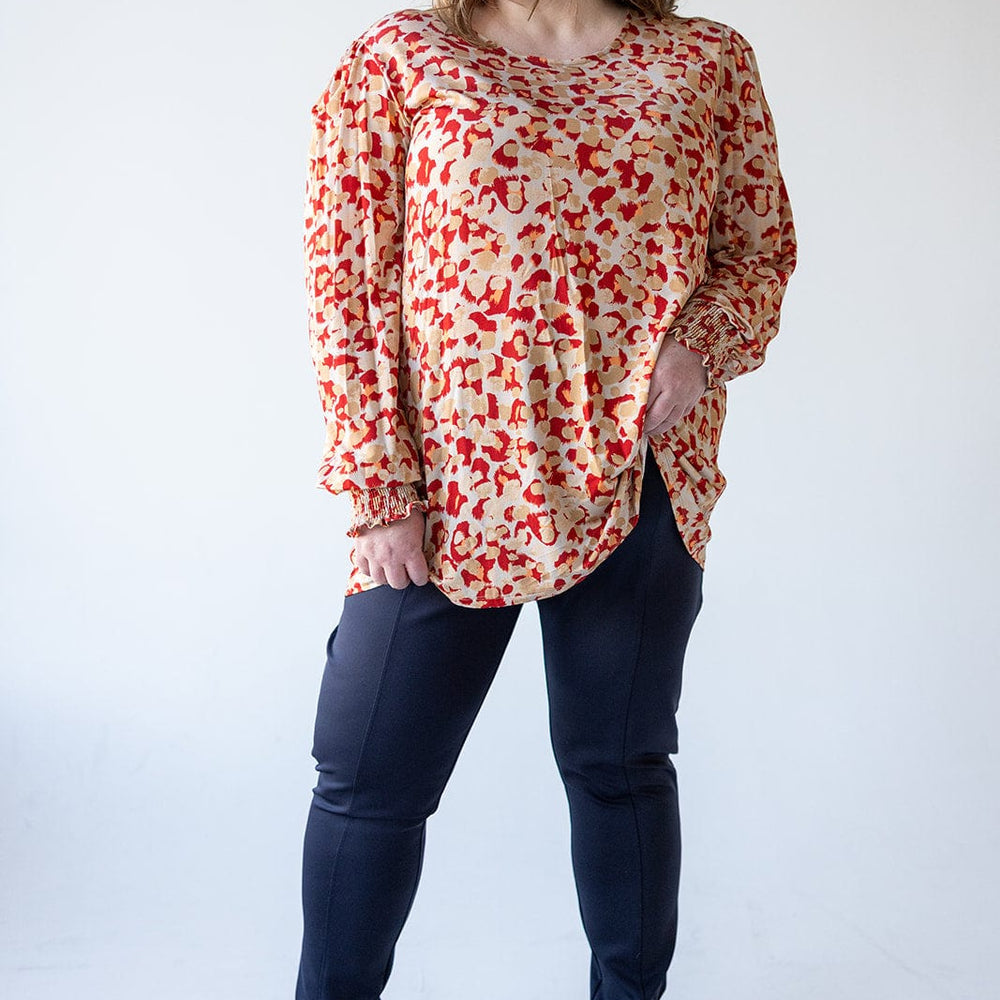 A person wearing a patterned blouse in sateen fabric, slim straight leg dark pants, and gray shoes stands smiling against a plain background.