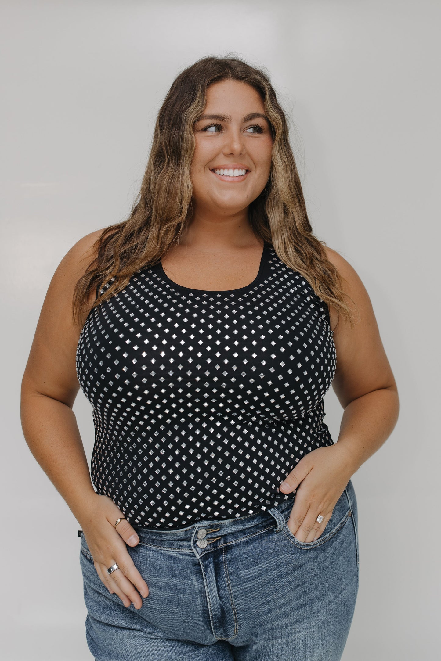Woman wearing a black tank top with white polka dots and blue jeans against a plain background