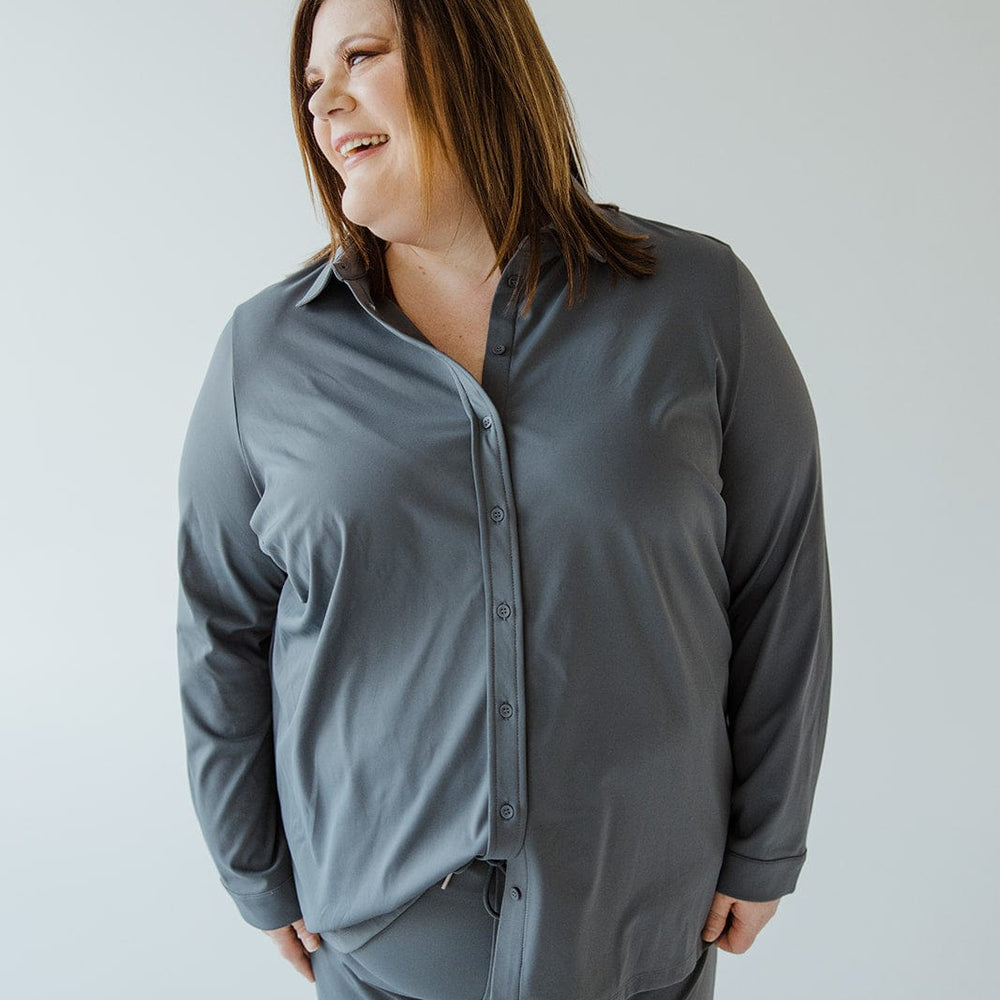 A person with shoulder-length hair wearing a gray Love Marlow STRETCHY WRINKLE FREE BUTTON-UP BLOUSE IN HAZY STEEL BLUE and matching pants smiles and looks to the side against a plain light-colored background.