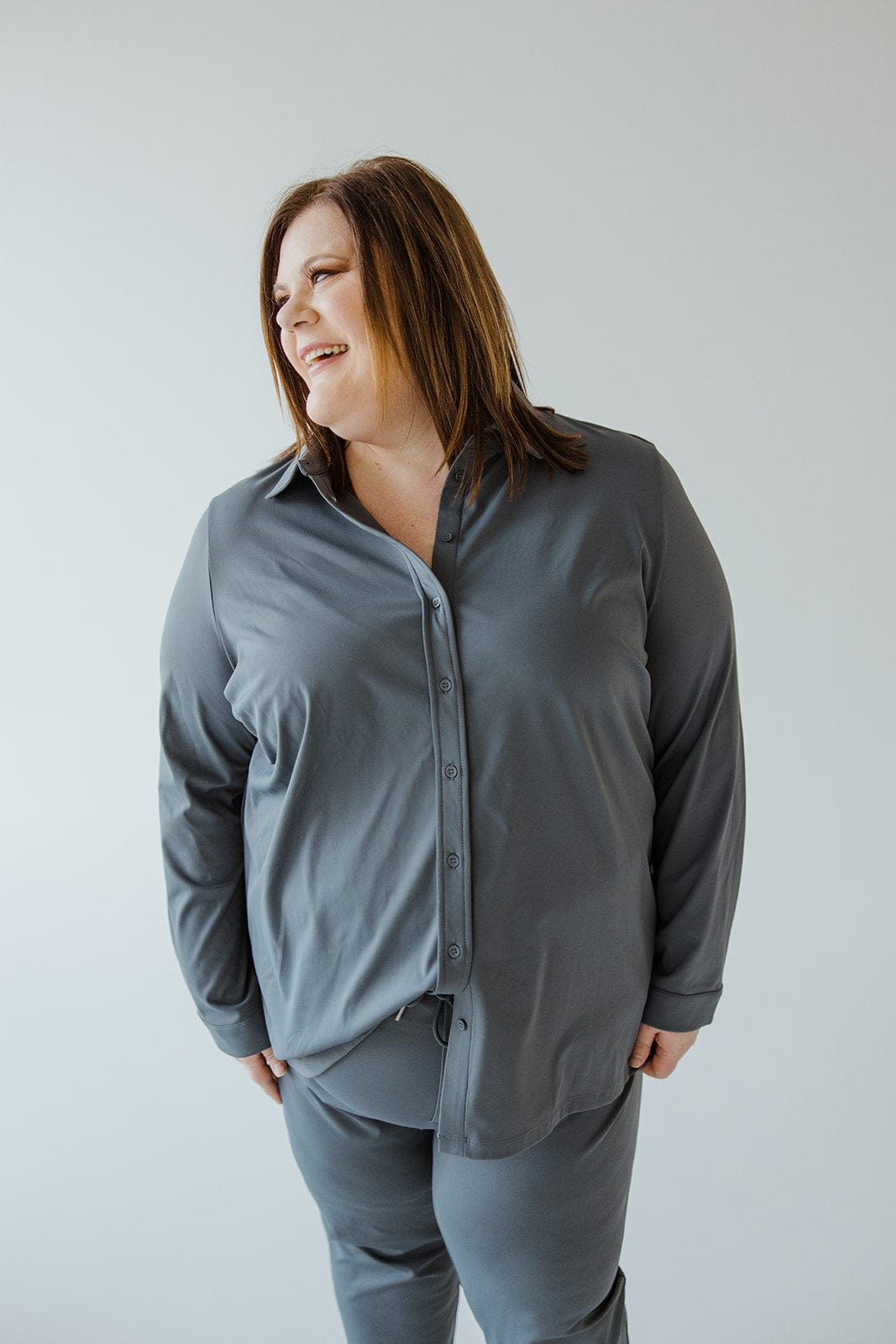 A person with shoulder-length hair wearing a gray Love Marlow STRETCHY WRINKLE FREE BUTTON-UP BLOUSE IN HAZY STEEL BLUE and matching pants smiles and looks to the side against a plain light-colored background.