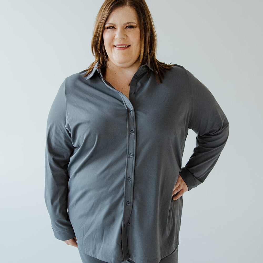 A woman stands with one hand on her hip, smiling. She is wearing a Love Marlow STRETCHY WRINKLE FREE BUTTON-UP BLOUSE IN HAZY STEEL BLUE and matching pants made of the same wrinkle-free fabric. The background is a simple, plain white.