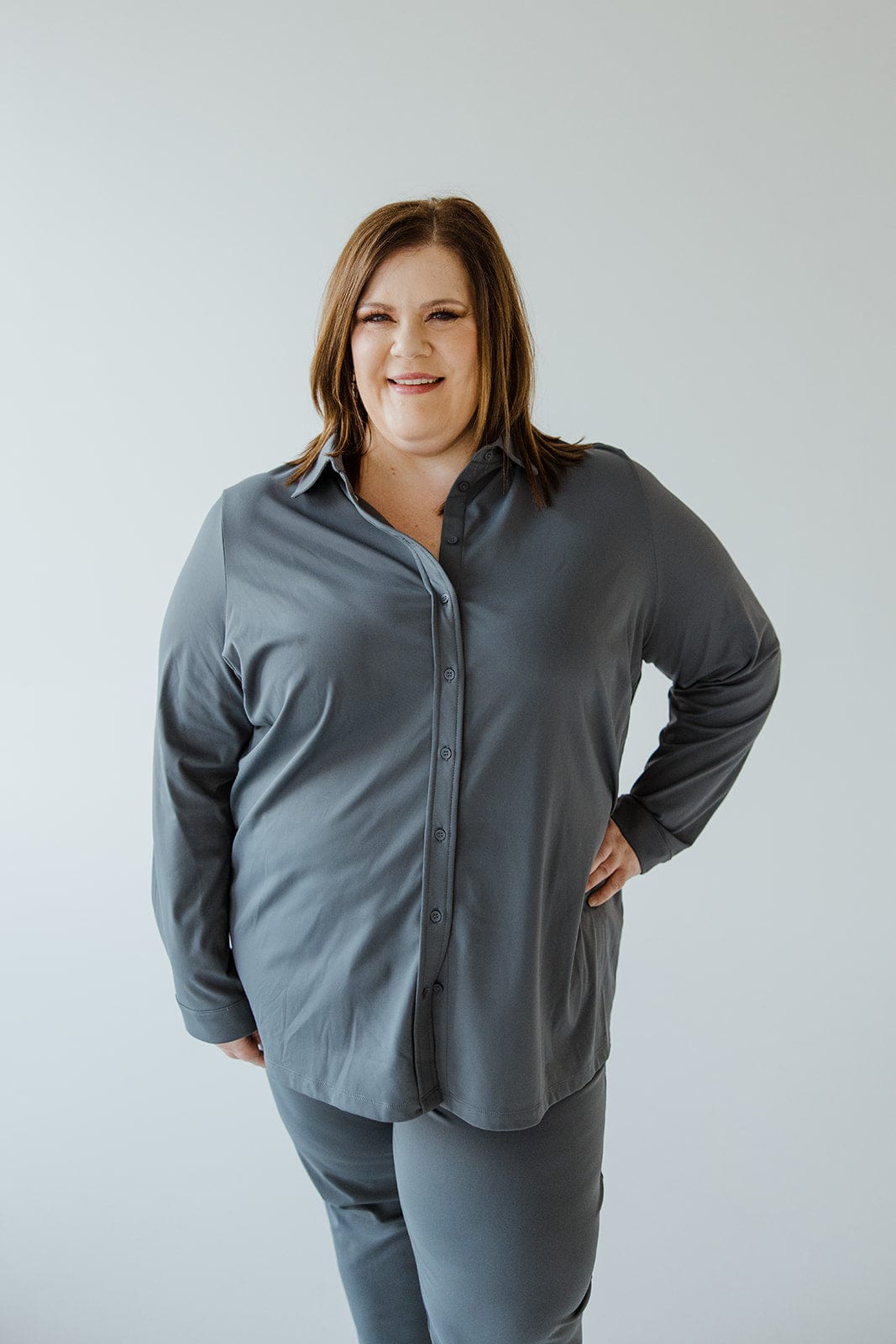 A woman stands with one hand on her hip, smiling. She is wearing a Love Marlow STRETCHY WRINKLE FREE BUTTON-UP BLOUSE IN HAZY STEEL BLUE and matching pants made of the same wrinkle-free fabric. The background is a simple, plain white.