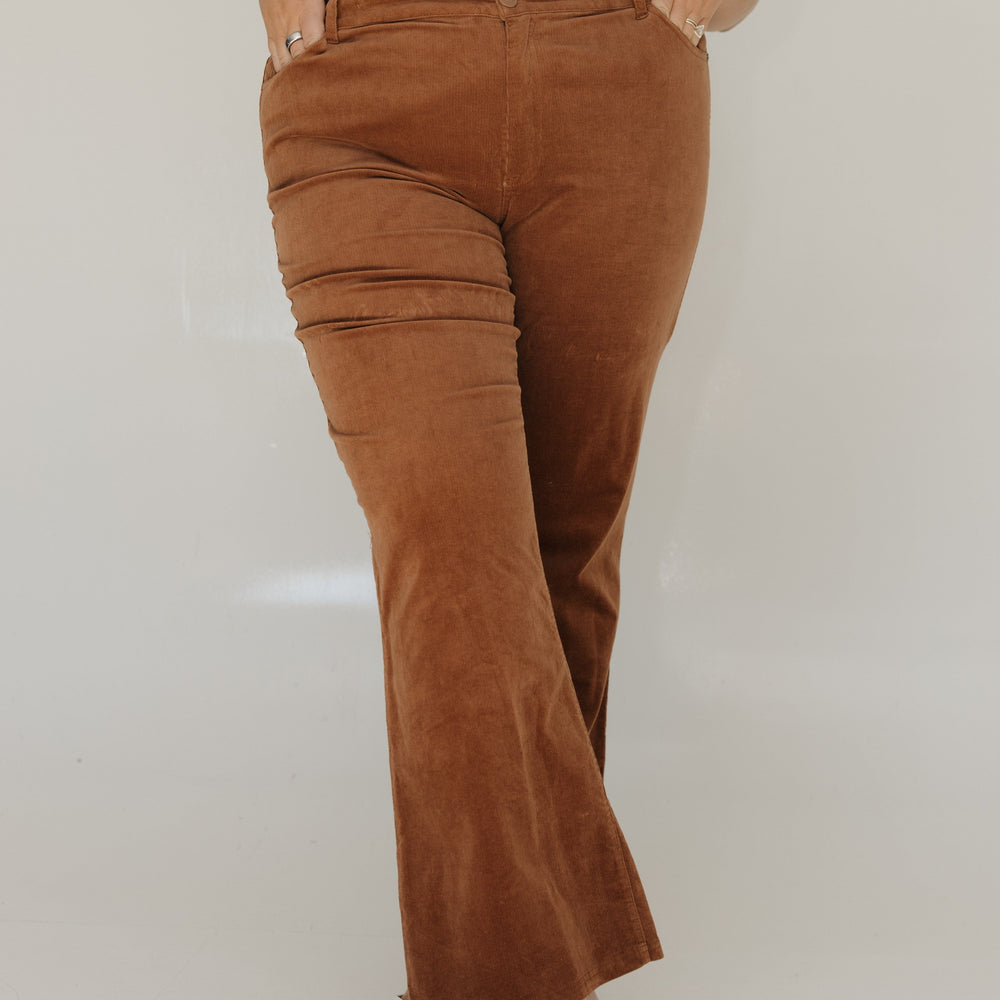 Person wearing brown flared pants and a gray t-shirt with a white graphic on a plain background