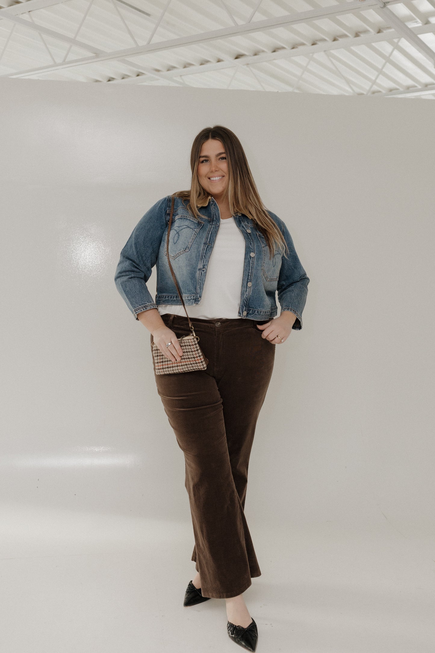Woman wearing a denim jacket, white top, and brown pants against a white background