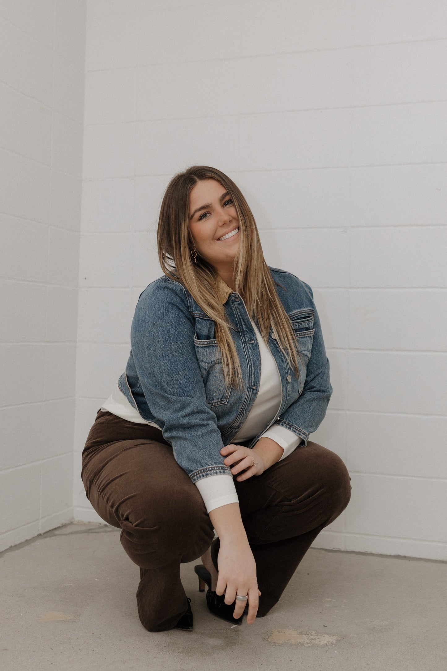 Woman in a denim jacket and brown pants sitting against a white brick wall.