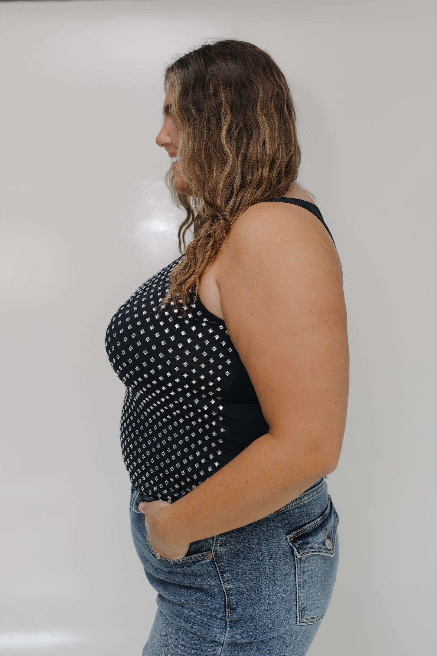 Woman wearing a black tank top with white polka dots and blue jeans against a plain background