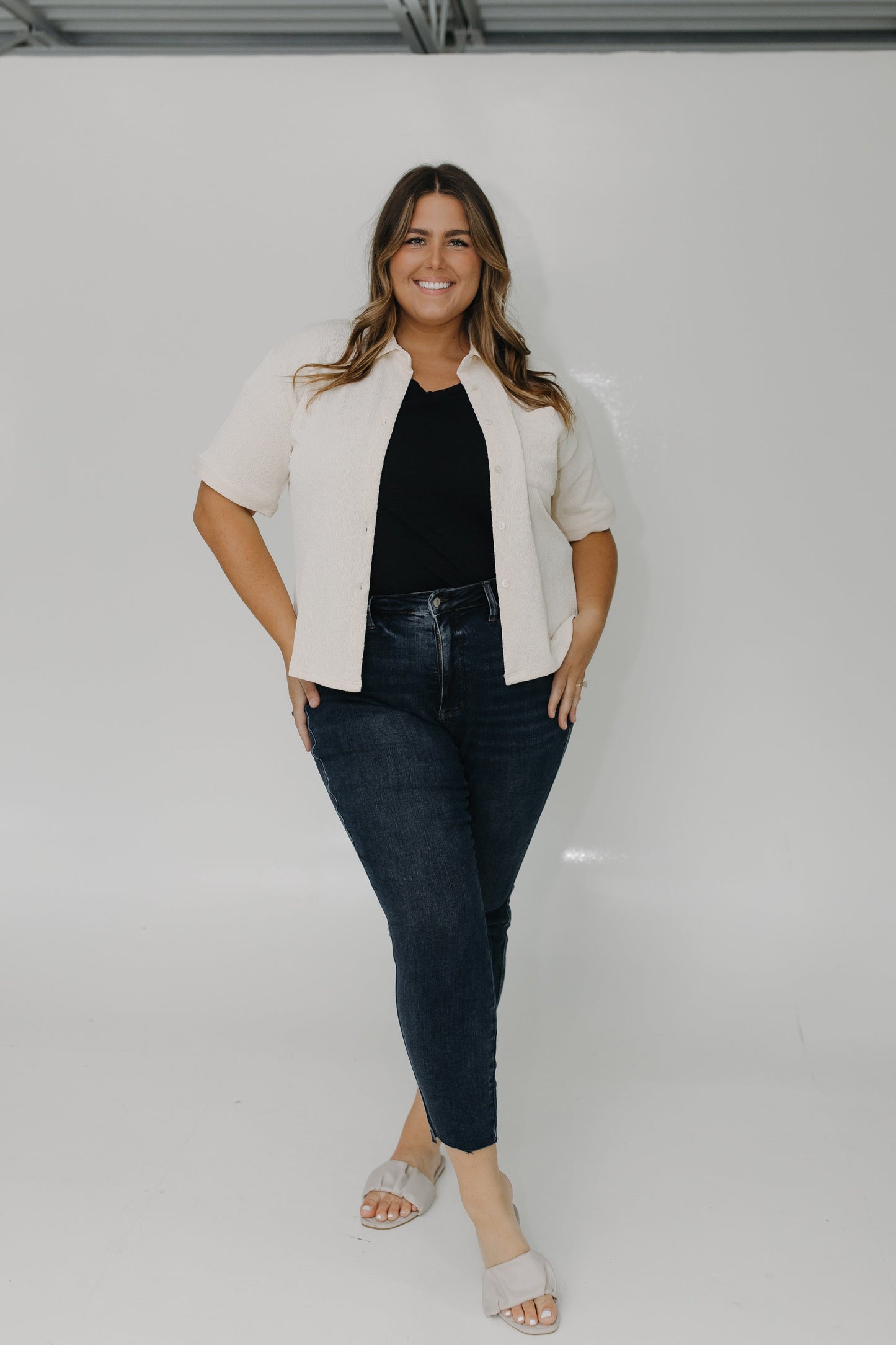 Woman wearing a white jacket, black top, and dark jeans against a plain background
