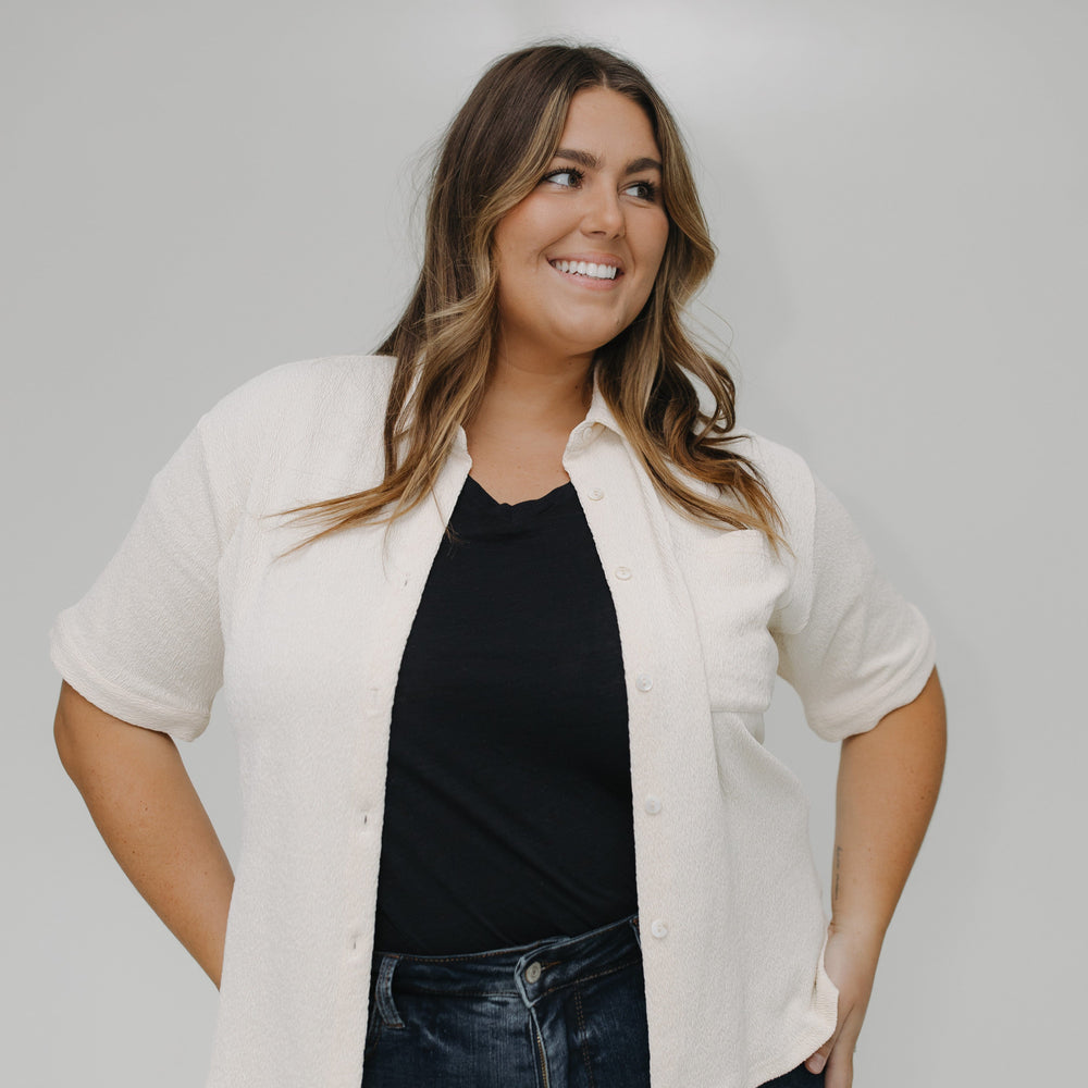 Woman wearing a white blouse over a black top and blue jeans against a plain background