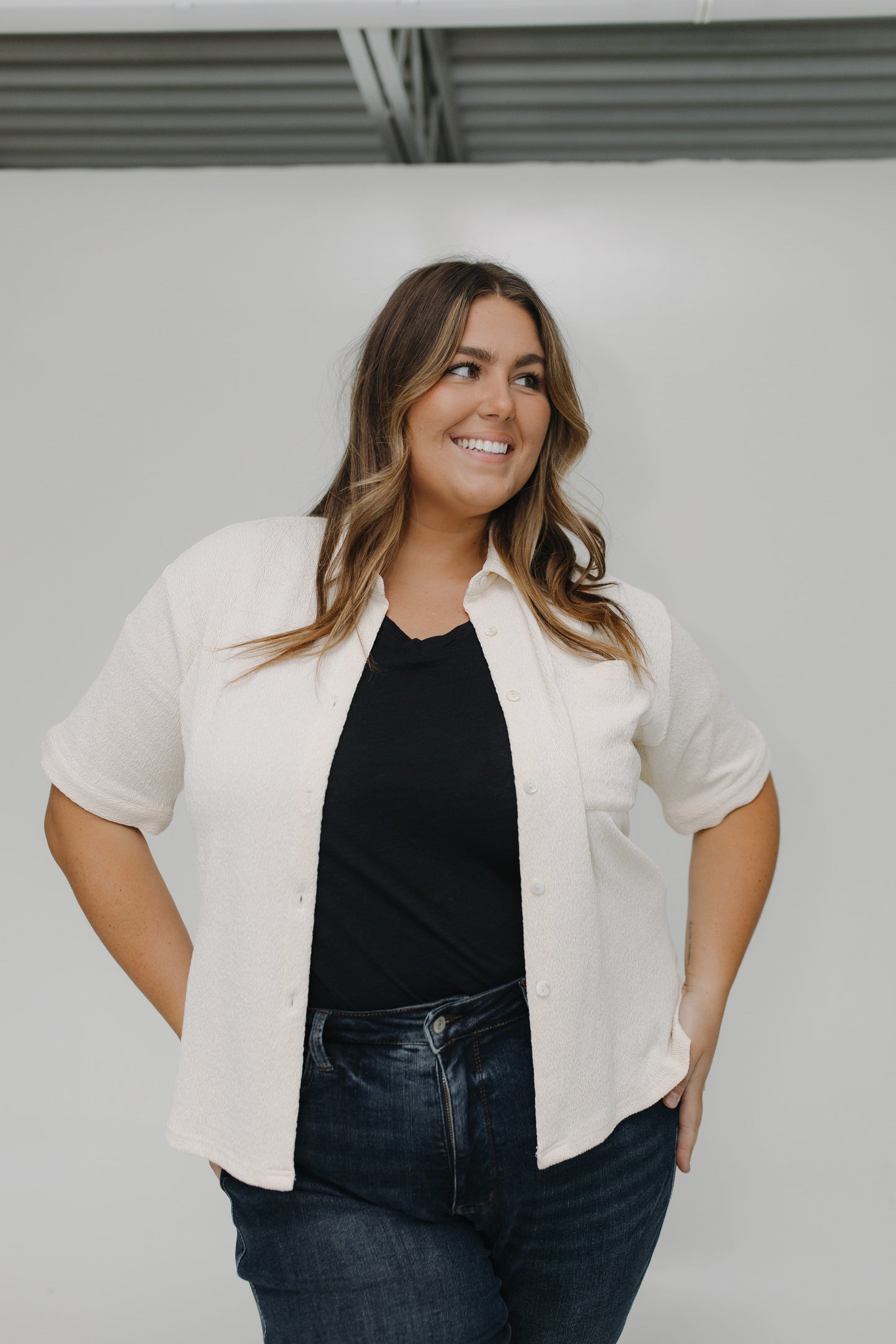 Woman wearing a white blouse over a black top and blue jeans against a plain background
