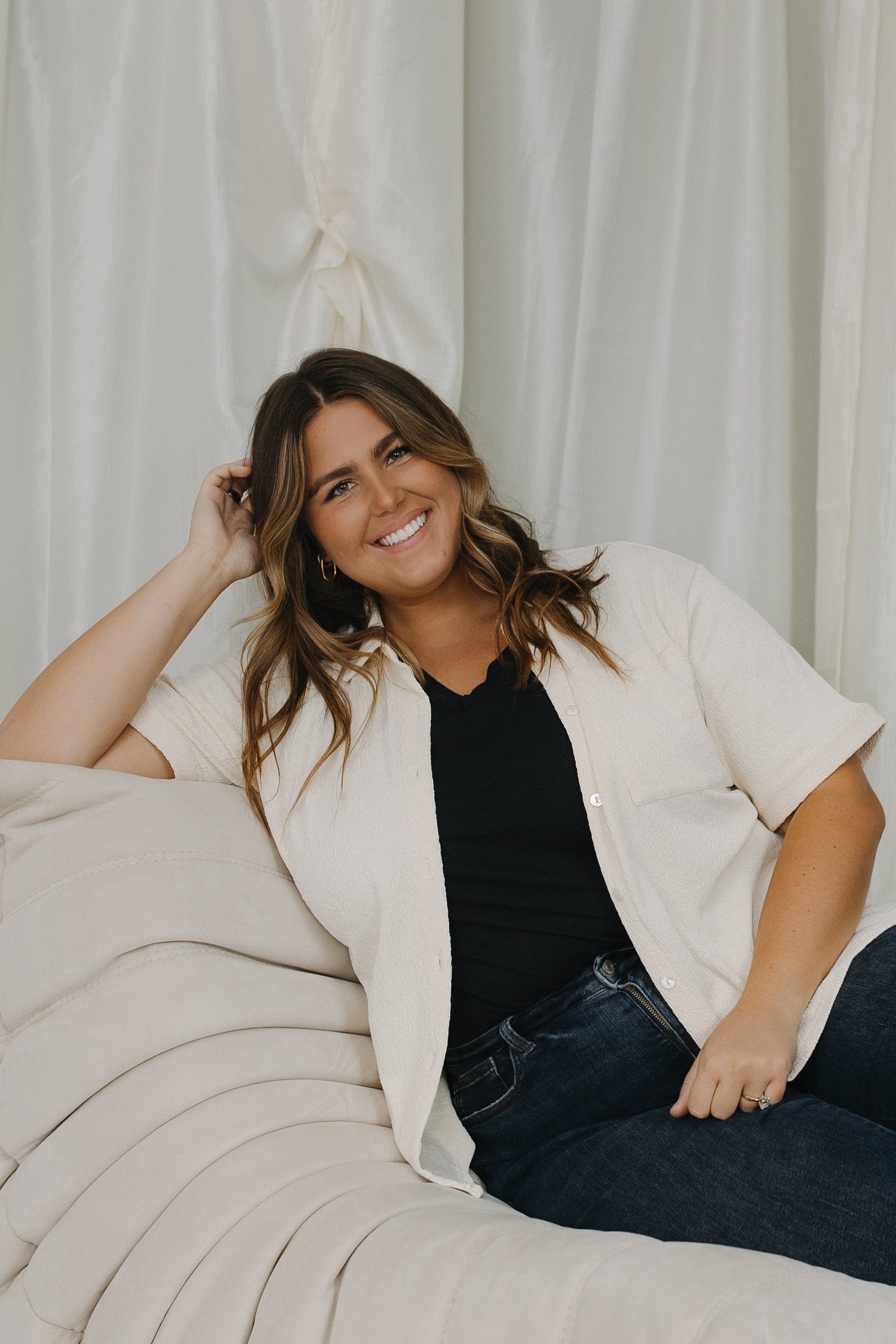 Woman sitting on a beige couch wearing a white cardigan, black top, and jeans against a white curtain background.