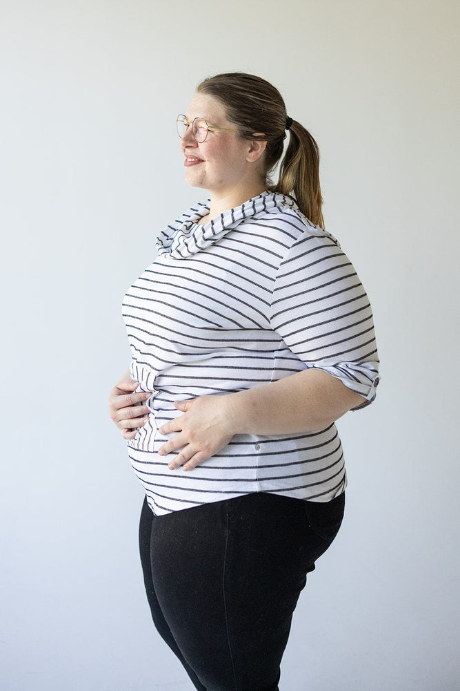 A person with glasses and a ponytail, wearing an off-white and black STRIPED COWL NECK HOODIE IN OFF-WHITE by Love Marlow and black pants, stands against a plain background, smiling and looking to the side.