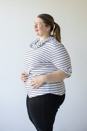 A person with glasses and a ponytail, wearing an off-white and black STRIPED COWL NECK HOODIE IN OFF-WHITE by Love Marlow and black pants, stands against a plain background, smiling and looking to the side.