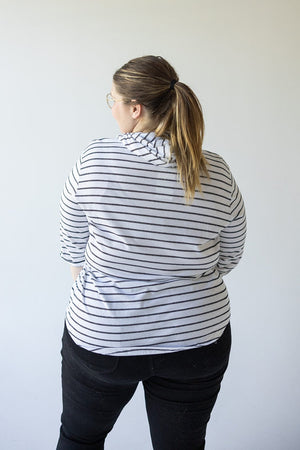 A person with long hair in a ponytail, wearing glasses, a Love Marlow STRIPED COWL NECK HOODIE IN OFF-WHITE with an off-white and black striped design, and black pants, facing away from the camera.