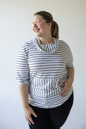 A woman wearing glasses and a Love Marlow STRIPED COWL NECK HOODIE IN OFF-WHITE smiles while looking to the side. She has her hair tied back and is standing against a plain background.