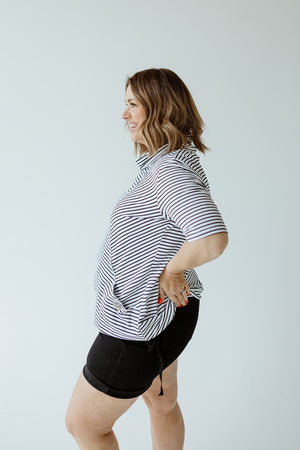 A woman with shoulder-length hair smiles, posing in a side profile view. She is wearing a Love Marlow STRIPED COWL NECK SHORT SLEEVE TUNIC IN OFF-WHITE and black shorts.