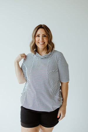 Person standing against a plain background, wearing a Love Marlow STRIPED COWL NECK SHORT SLEEVE TUNIC IN OFF-WHITE and black shorts, smiling, with one hand adjusting the collar of the tunic.