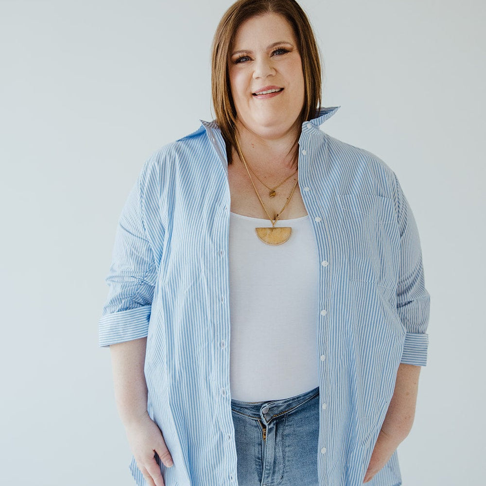 A person stands against a plain background, wearing the Love Marlow STRIPED TUNIC BUTTON-UP BLOUSE IN OFF-WHITE AND AZURE over a white top, paired with blue jeans.