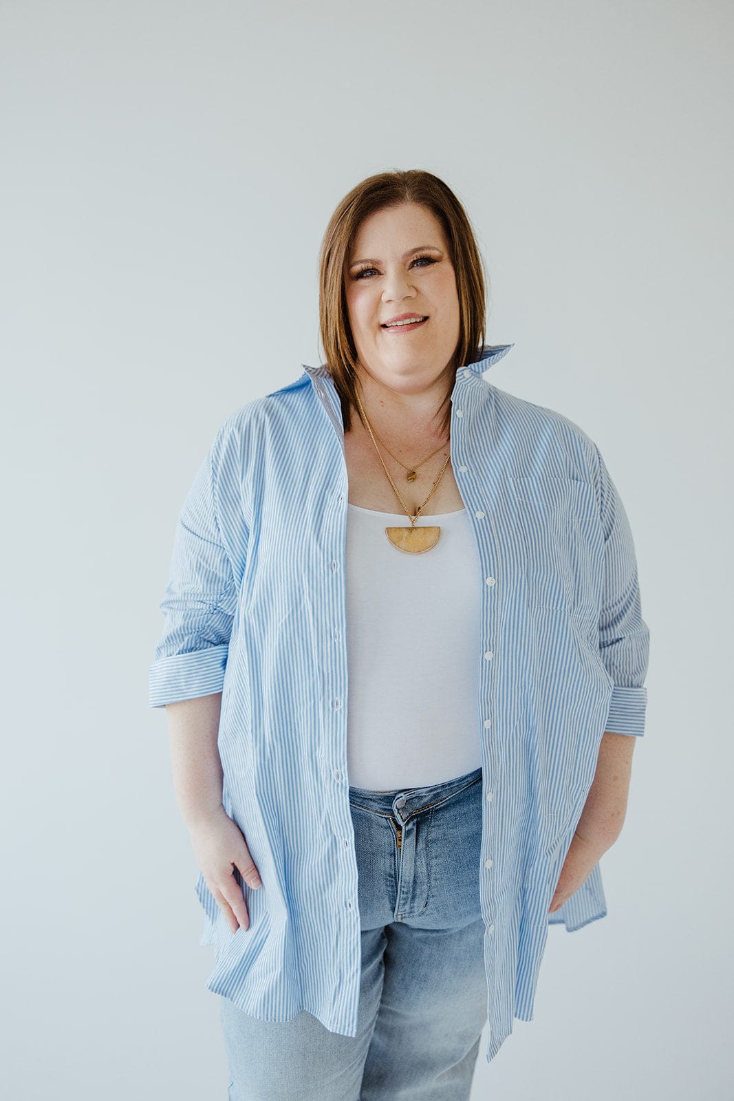 A person stands against a plain background, wearing the Love Marlow STRIPED TUNIC BUTTON-UP BLOUSE IN OFF-WHITE AND AZURE over a white top, paired with blue jeans.