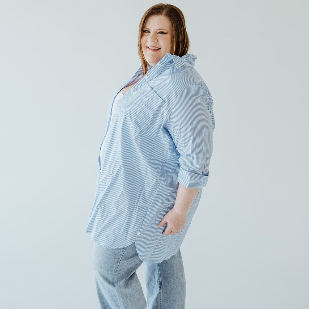 A person smiles at the camera while standing against a plain background, dressed in light blue jeans and the "Striped Tunic Button-Up Blouse in Off-White and Azure" by Love Marlow.