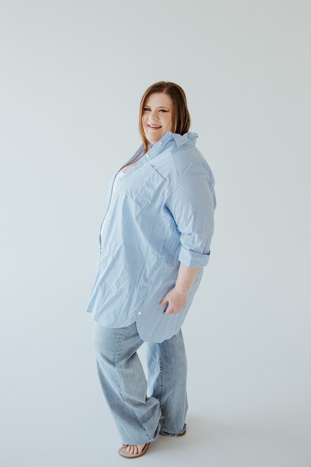 A person smiles at the camera while standing against a plain background, dressed in light blue jeans and the "Striped Tunic Button-Up Blouse in Off-White and Azure" by Love Marlow.