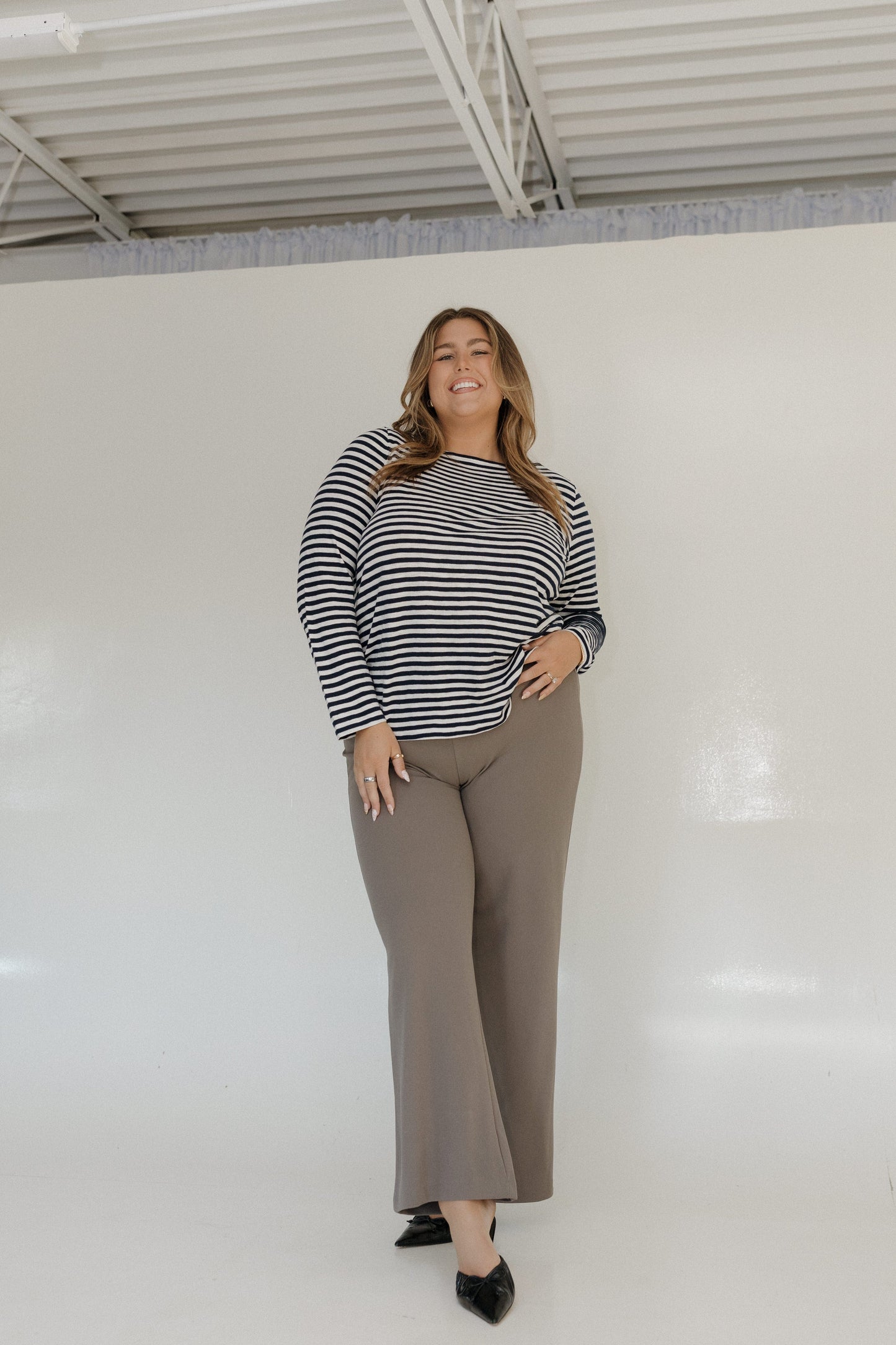 Woman wearing a striped sweater and beige pants standing against a white wall.
