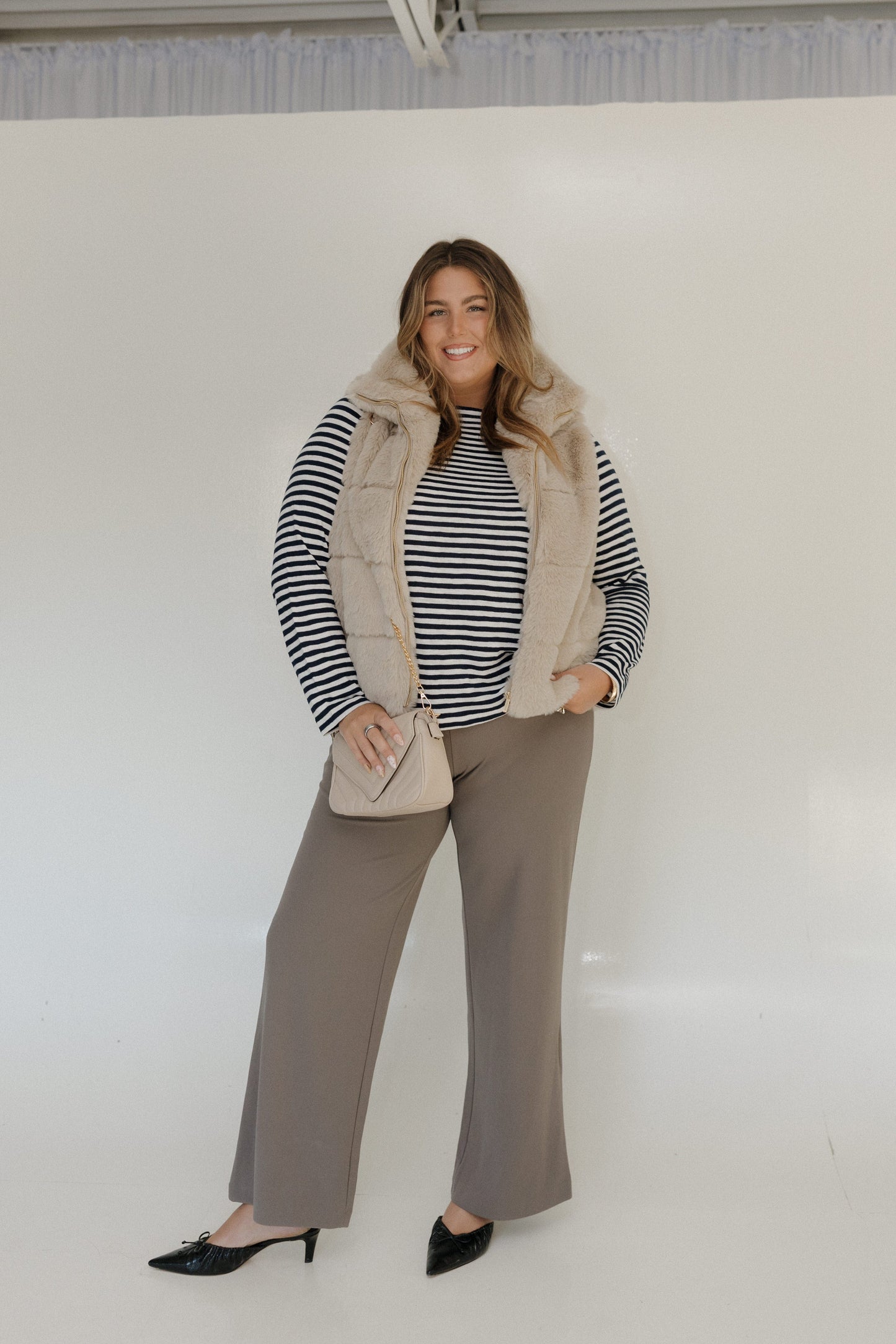 Woman wearing a striped sweater, beige vest, and gray pants against a white background