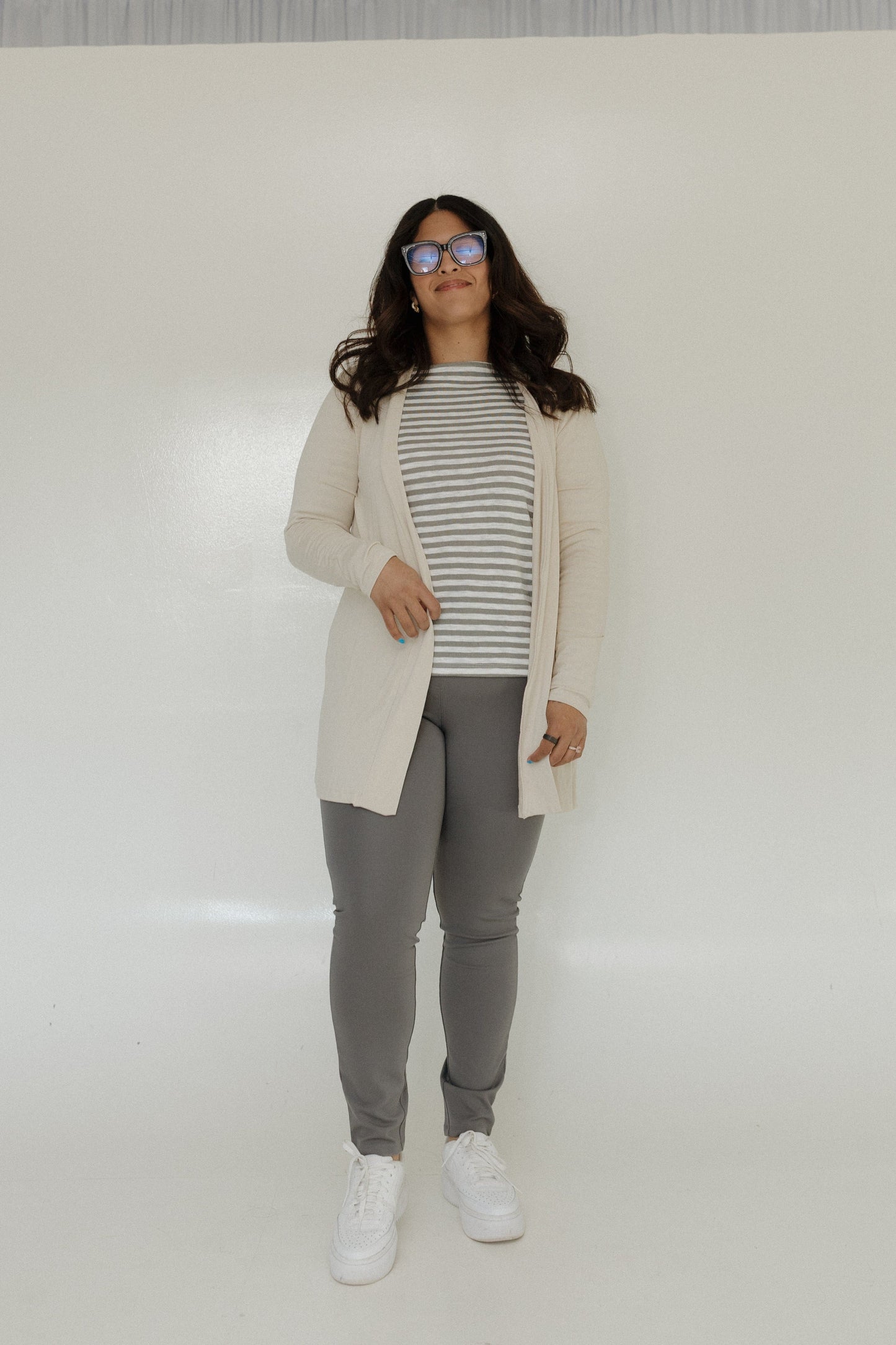 Person wearing a beige coat, striped shirt, gray pants, and white sneakers on a white background