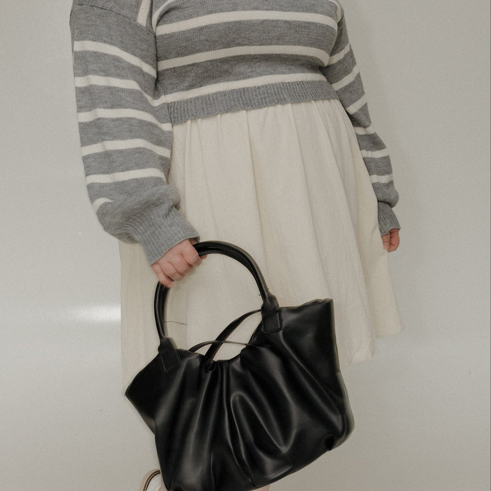 Woman wearing a gray striped sweater and white skirt, holding a black handbag against a plain background