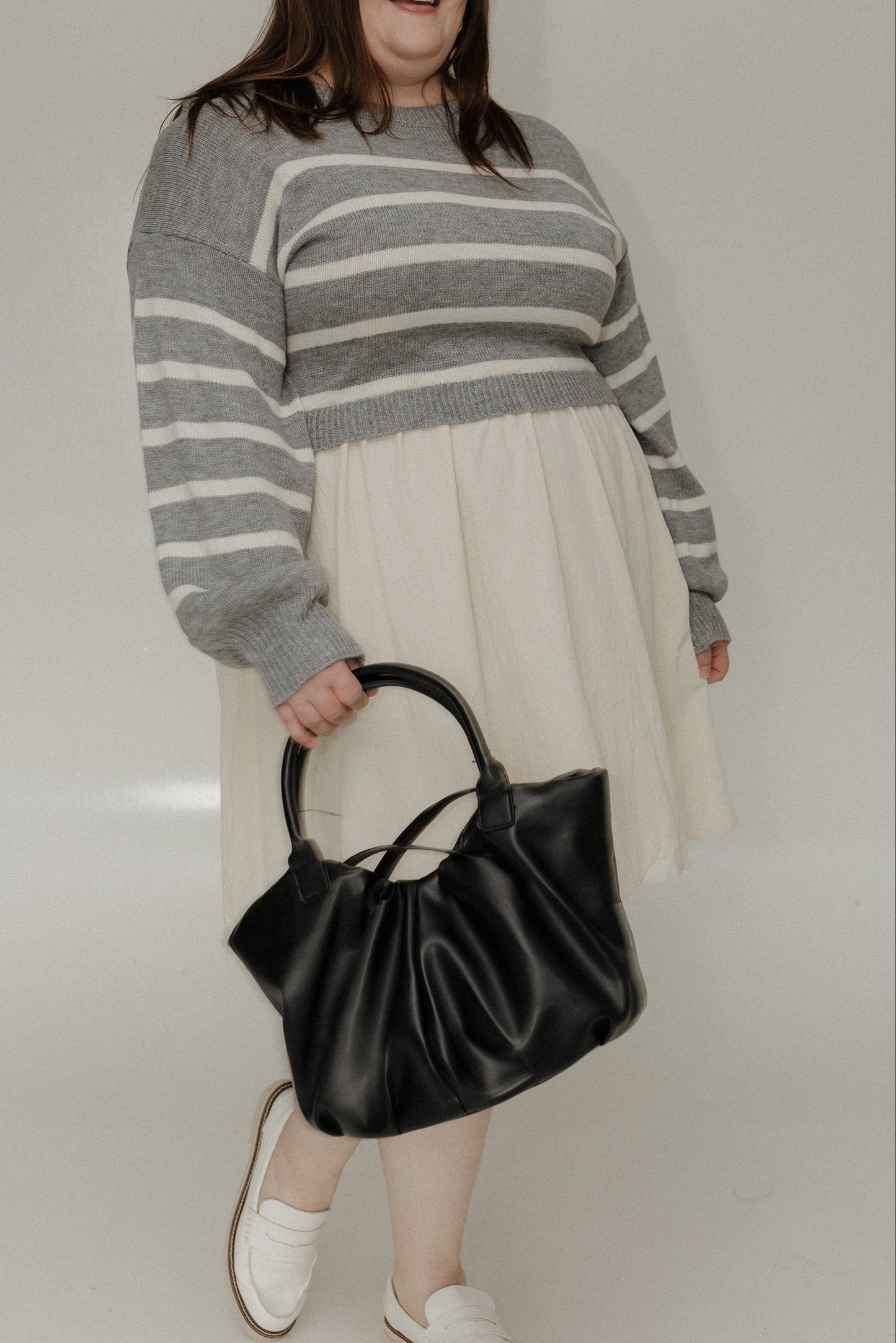 Woman wearing a gray striped sweater and white skirt, holding a black handbag against a plain background