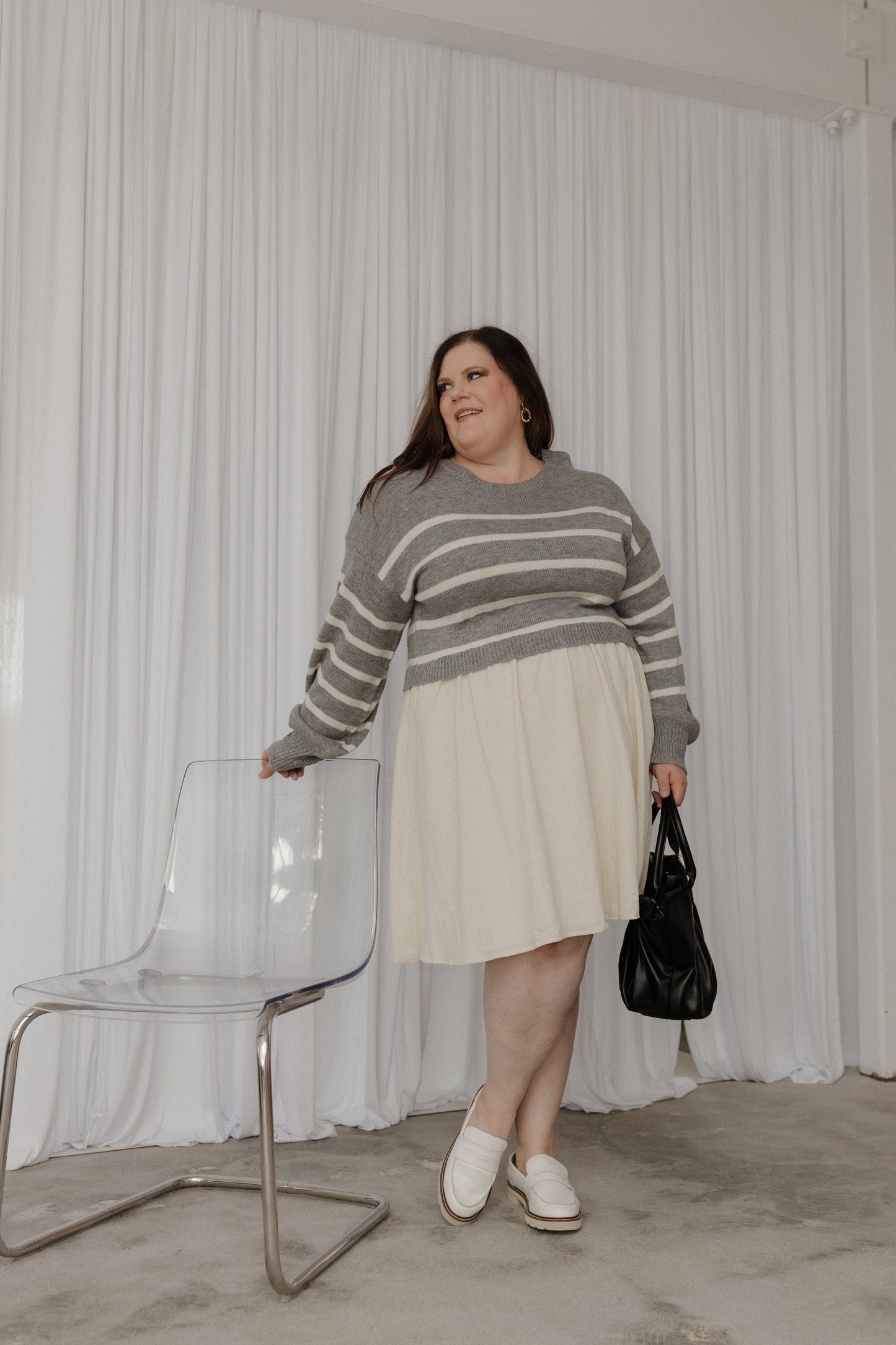 Woman in a striped sweater and skirt standing next to a clear chair against a white curtain.