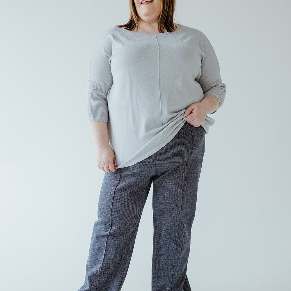 A person stands against a plain background, wearing a light sweater crafted from luxurious knit fabric and gray high-waist pants, holding the edge of the sweater with one hand.