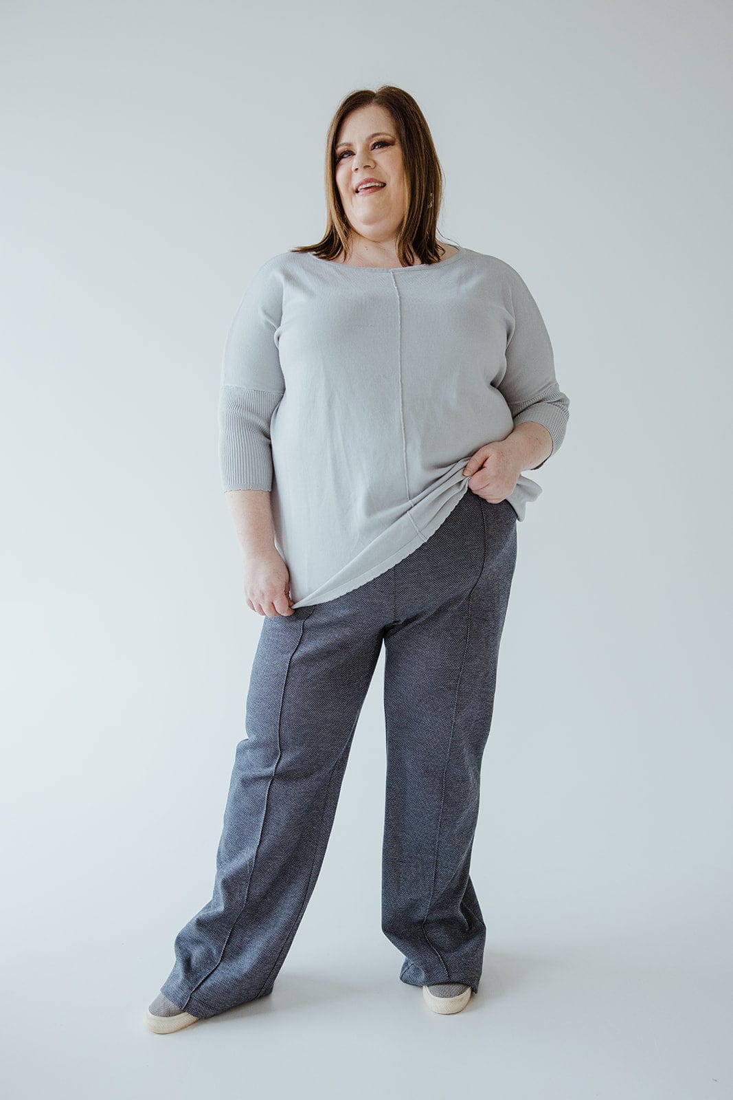 A person stands against a plain background, wearing a light sweater crafted from luxurious knit fabric and gray high-waist pants, holding the edge of the sweater with one hand.