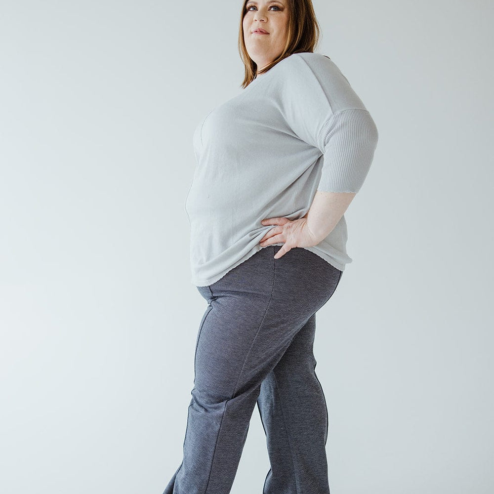 A person wearing a gray sweater crafted from luxurious knit fabric and high-waist blue pants stands with one hand on their hip, exuding confidence and professionalism against a plain background.