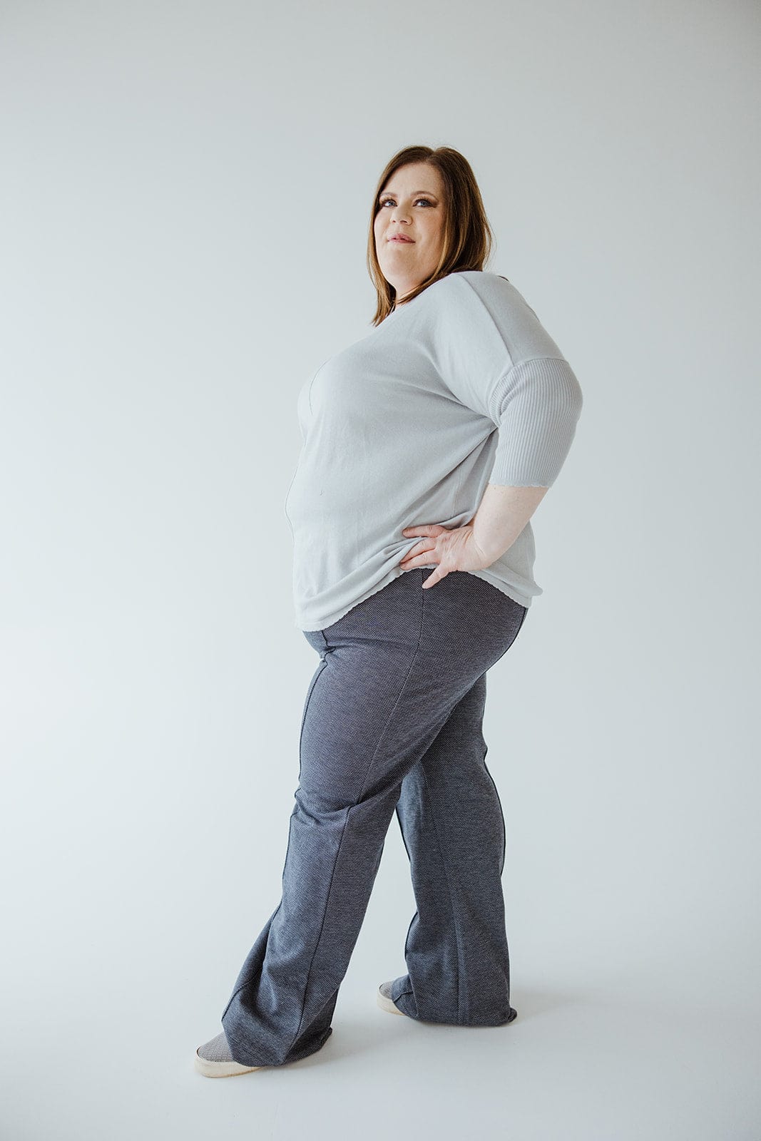 A person wearing a gray sweater crafted from luxurious knit fabric and high-waist blue pants stands with one hand on their hip, exuding confidence and professionalism against a plain background.