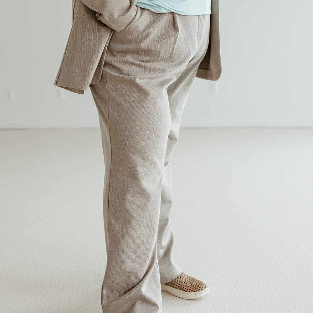 A person in a beige suit and light blue shirt with a subtle stripe pattern stands with one hand in their pocket, showcasing a sophisticated style. They are wearing woven slip-on shoes.