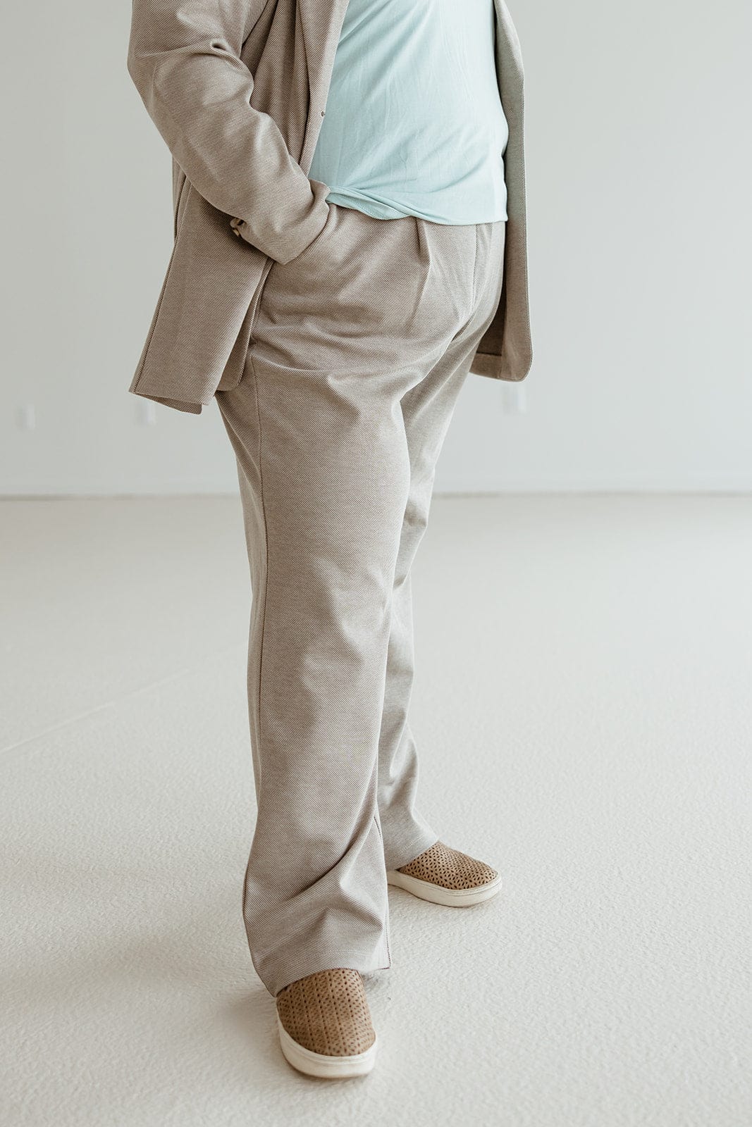 A person in a beige suit and light blue shirt with a subtle stripe pattern stands with one hand in their pocket, showcasing a sophisticated style. They are wearing woven slip-on shoes.