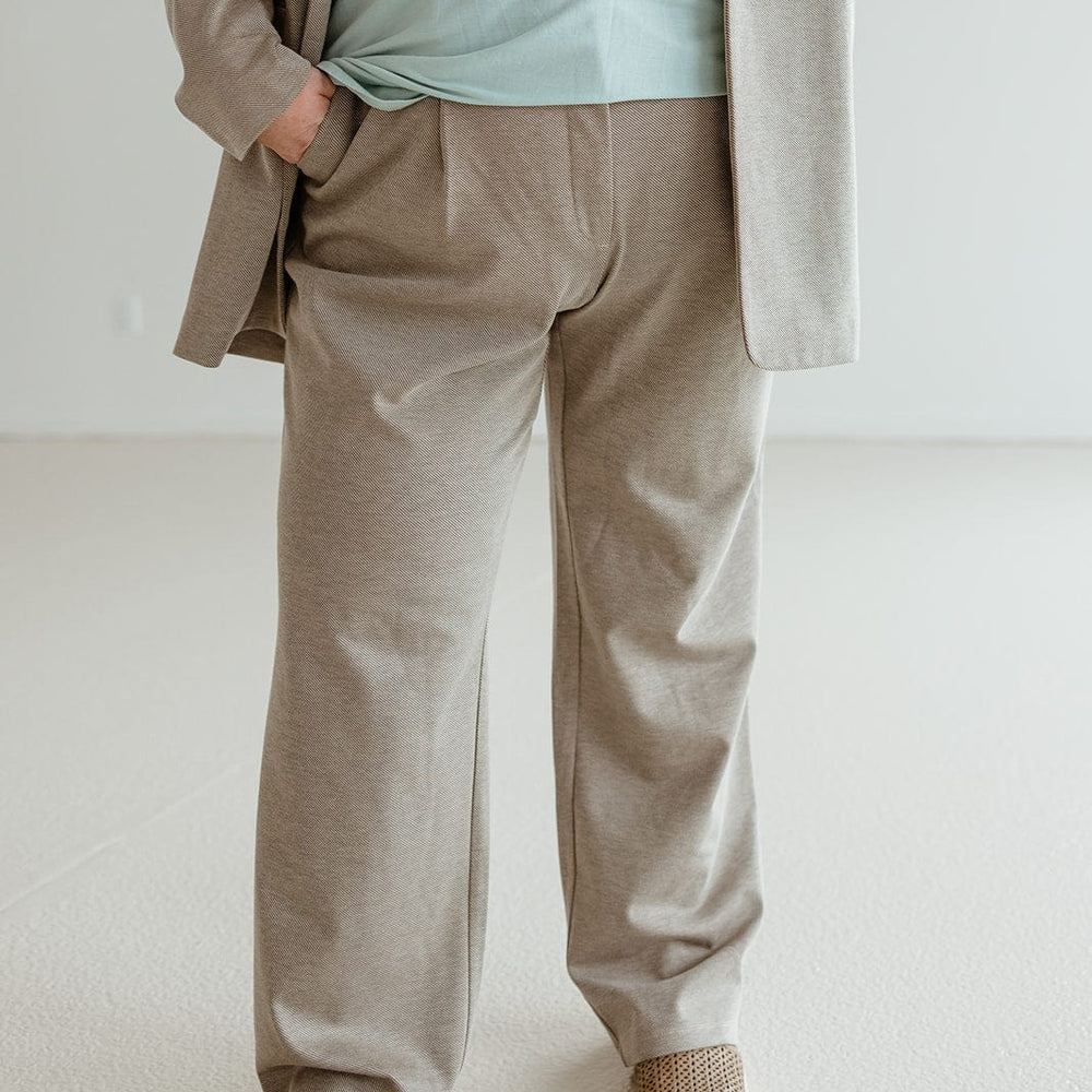 Person sporting a sophisticated style in a beige suit and green shirt, hands in pockets, standing in a light room. The ensemble is completed with brown slip-on shoes paired elegantly with jacquard woven trousers.