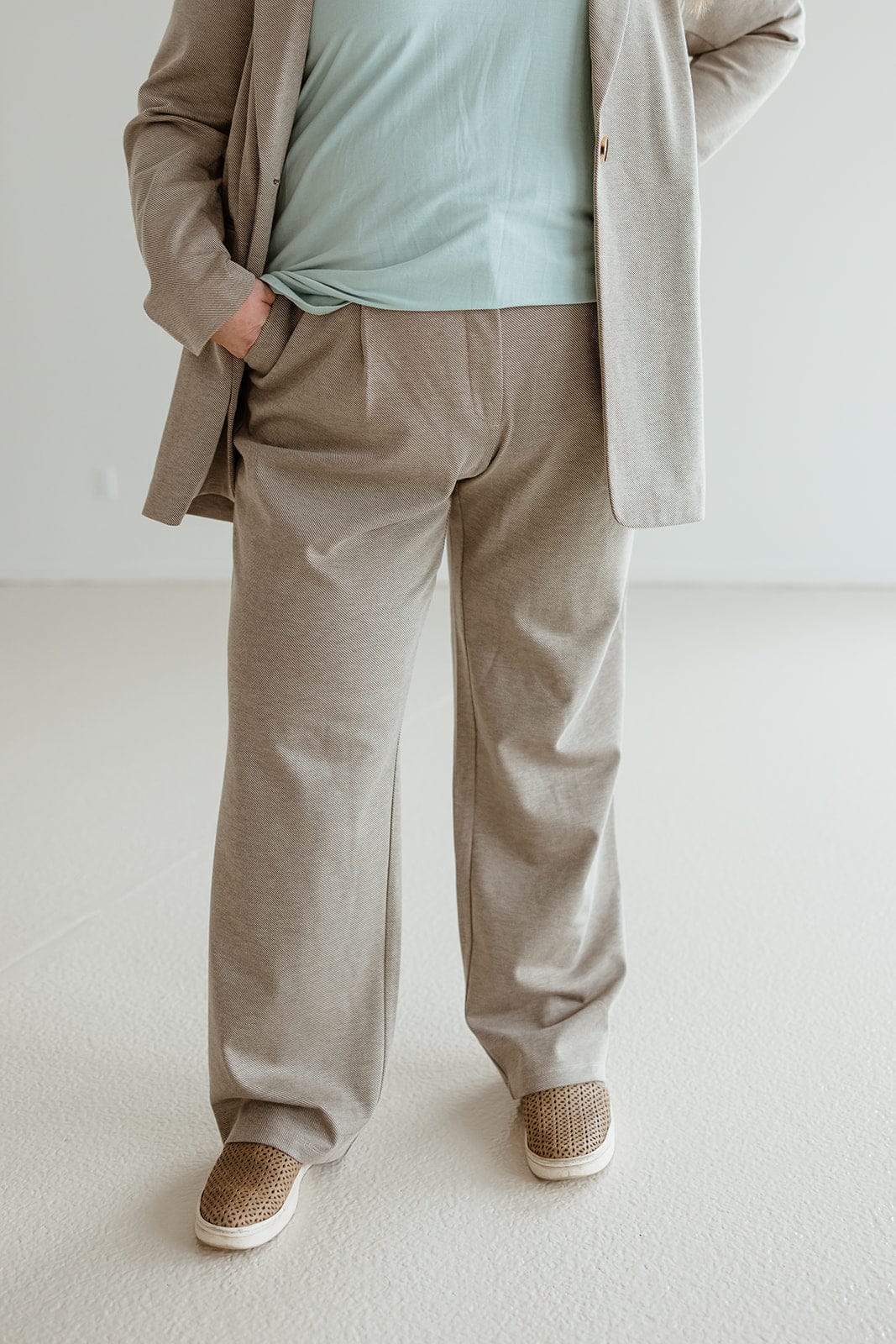 Person sporting a sophisticated style in a beige suit and green shirt, hands in pockets, standing in a light room. The ensemble is completed with brown slip-on shoes paired elegantly with jacquard woven trousers.