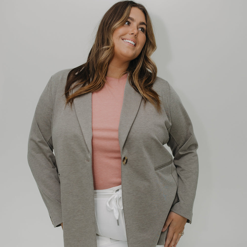 Woman wearing a gray blazer over a pink top and white pants against a white background