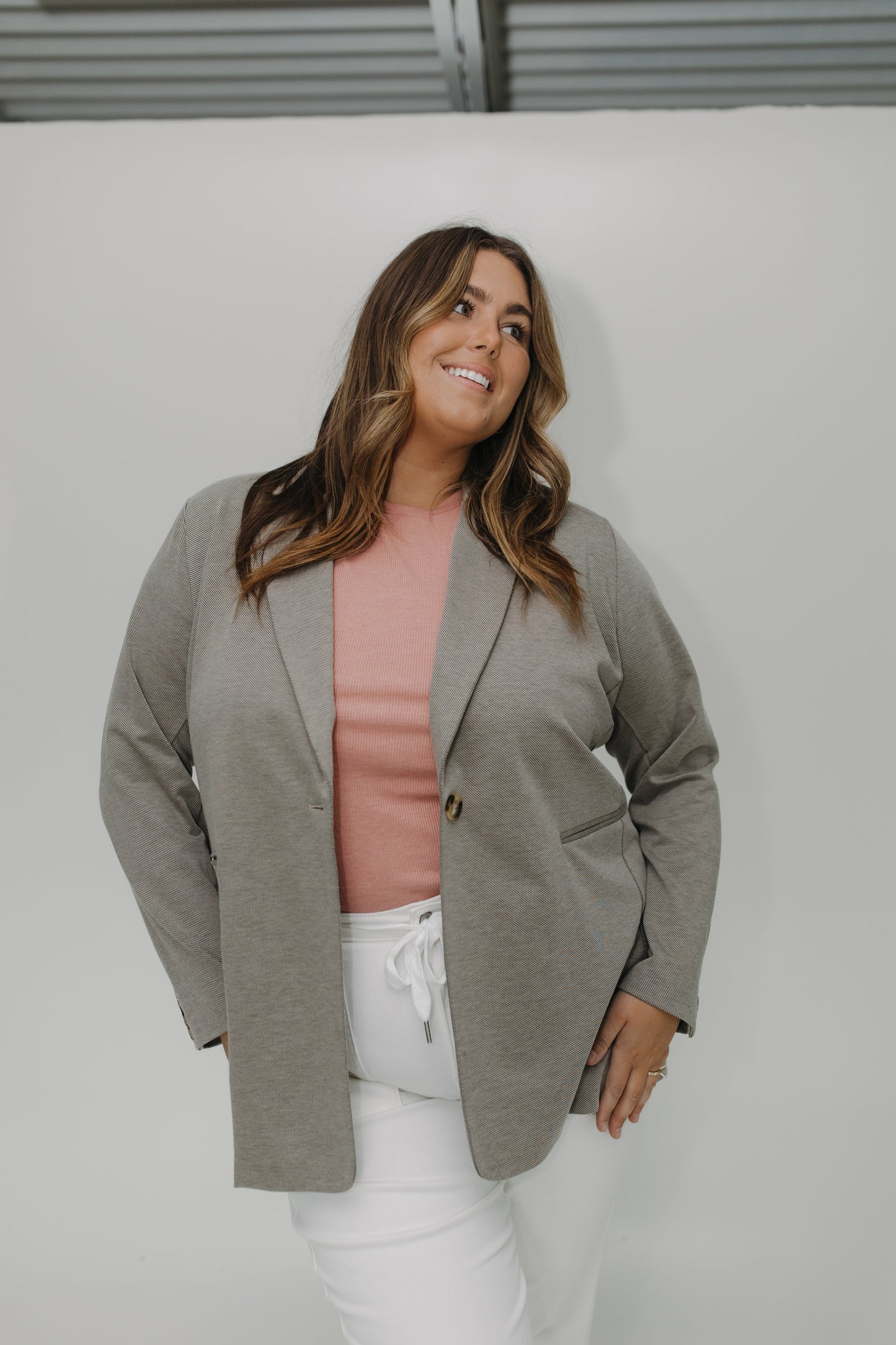 Woman wearing a gray blazer over a pink top and white pants against a white background