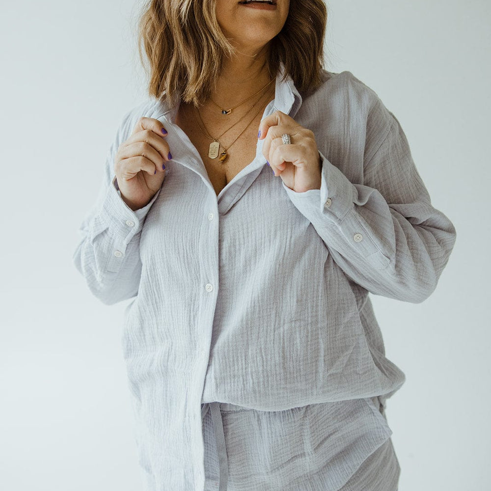 A woman with wavy hair wearing a Love Marlow SUPER SOFT TEXTURED BUTTON-UP BLOUSE in a light color, paired with shorts, is looking to her left against a plain white background.